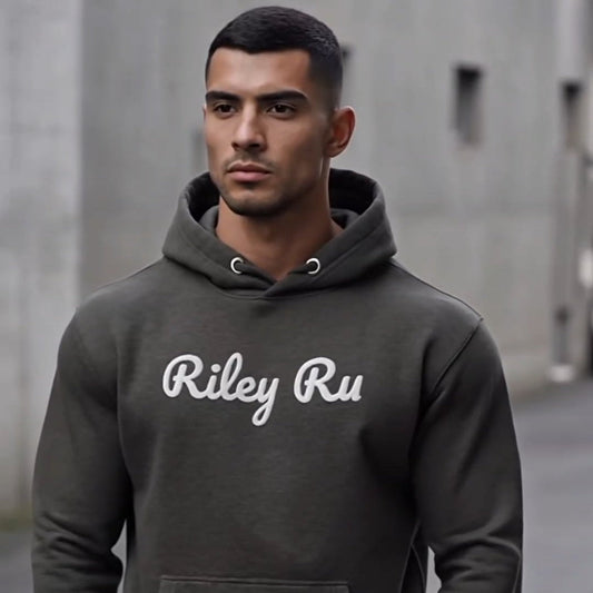 Man wearing a dark gray hoodie with 'Riley Ru' text, standing against a concrete wall.