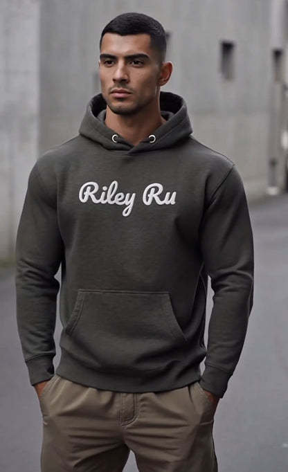 Man wearing a dark gray hoodie with 'Riley Ru' text, standing against a concrete wall.