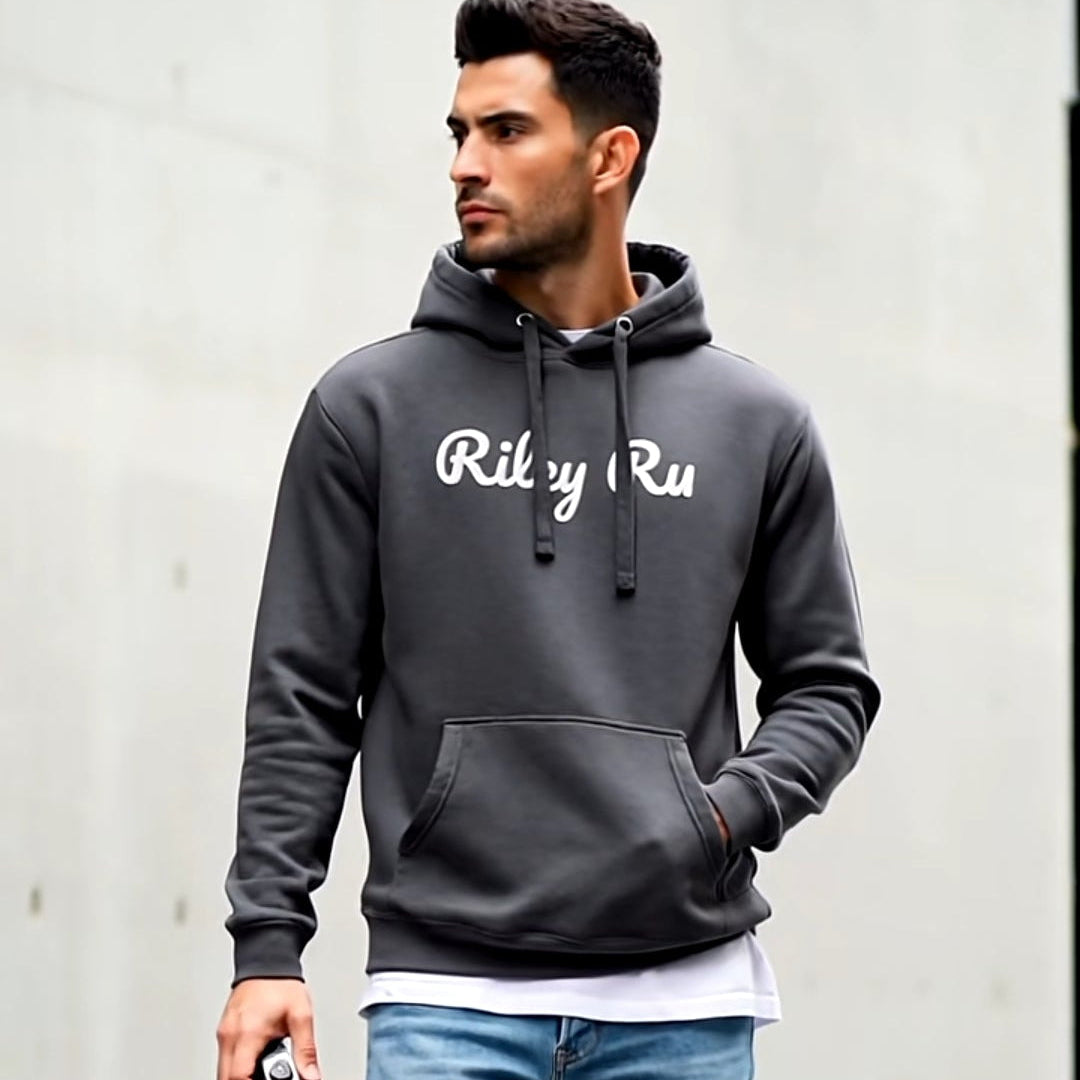 Man wearing a dark gray hoodie with 'Ricky Ru' text, light blue jeans, and white sneakers, standing against a plain wall.