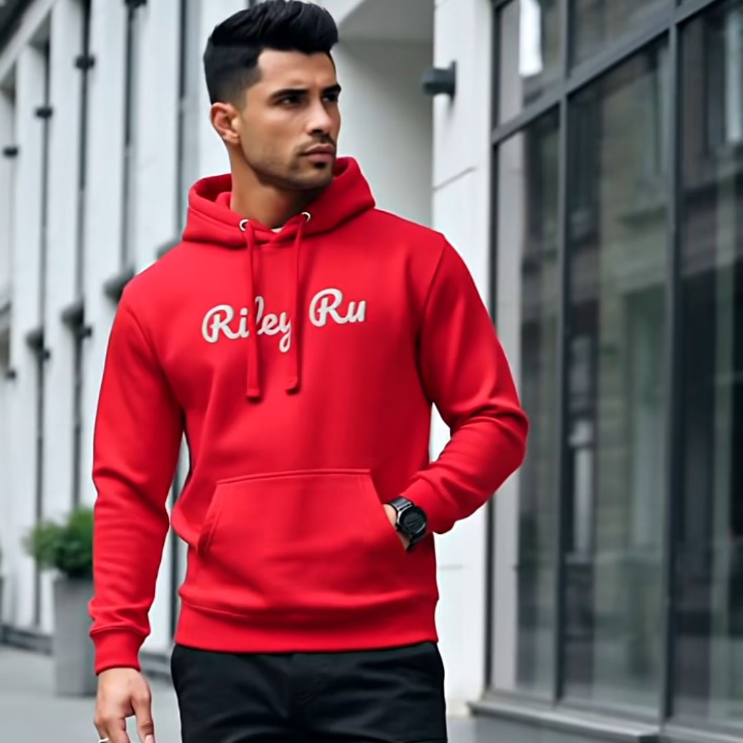 Man wearing a red hoodie with 'Riley Rue' branding, black pants, and white sneakers on a city street.