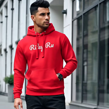 Man wearing a red hoodie with 'Riley Rue' branding, black pants, and white sneakers on a city street.