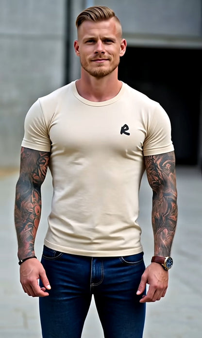 Man wearing a beige t-shirt with a logo and blue jeans standing against a gray wall.
