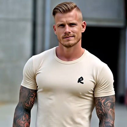 Man wearing a beige t-shirt and blue jeans with tattoos on arms, standing outdoors.