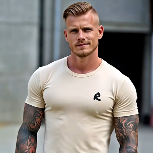 Man wearing a beige t-shirt and blue jeans with tattoos on arms, standing outdoors.