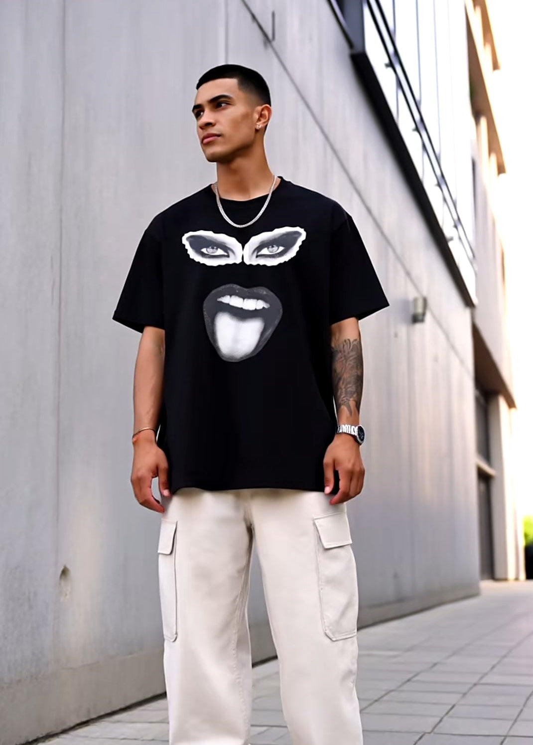Person wearing a black t-shirt with a graphic design and light-colored pants standing on a sidewalk.
