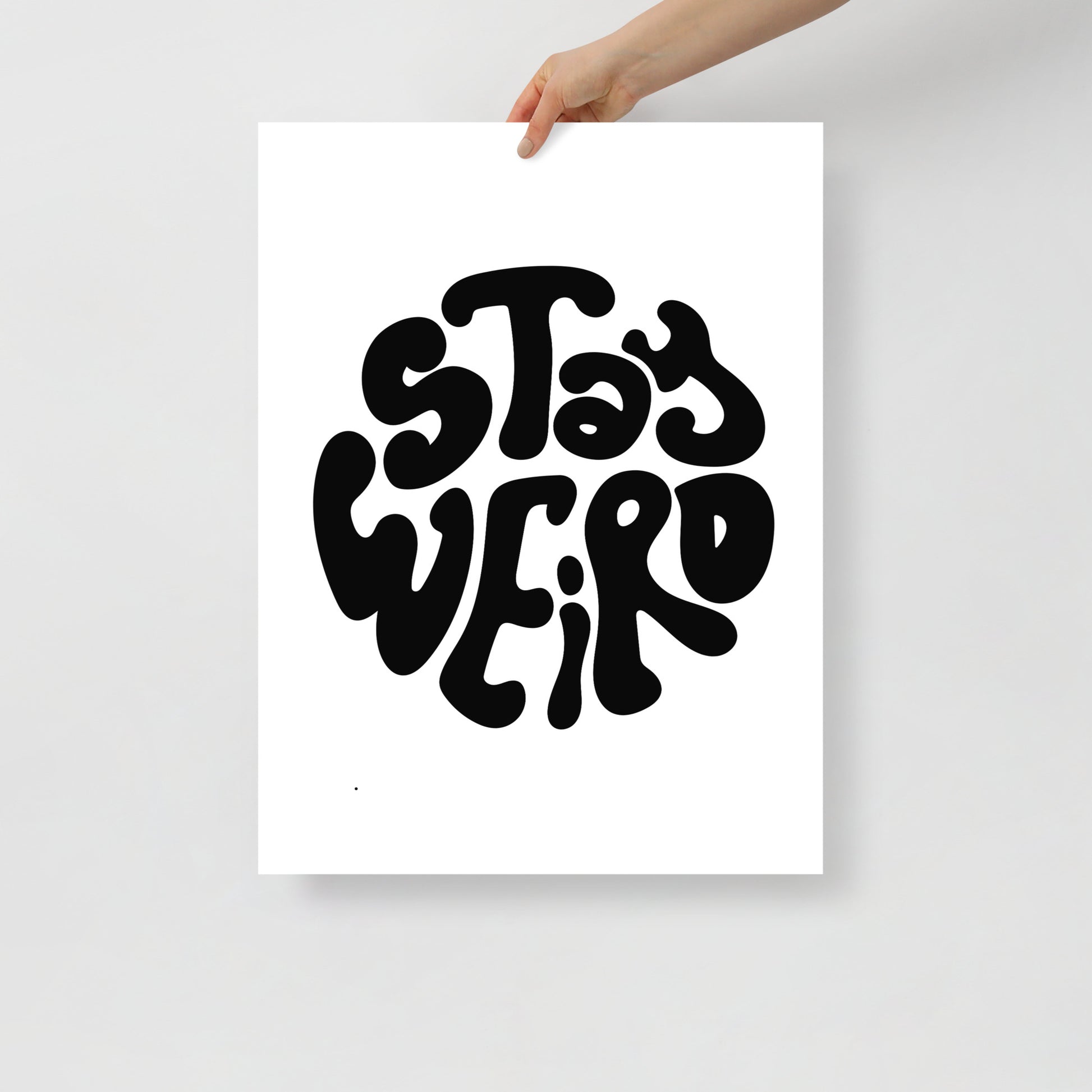Poster with 'Stay Weird' text held by a hand on a light gray background