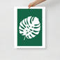 Framed poster with a white leaf design on a green background, held by a hand against a light gray background.