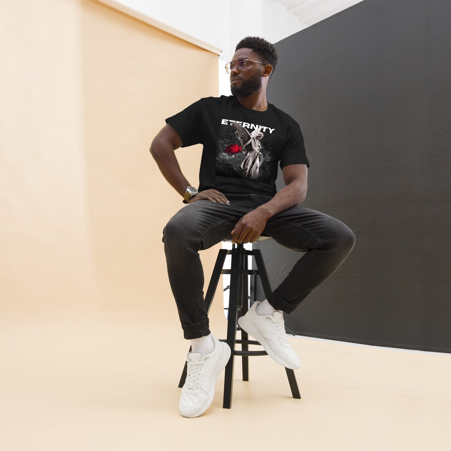 Man sitting on a stool wearing a black t-shirt with a graphic design, dark pants, and white sneakers.