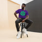Man sitting on a stool wearing a purple t-shirt with green text, black pants, and white sneakers.