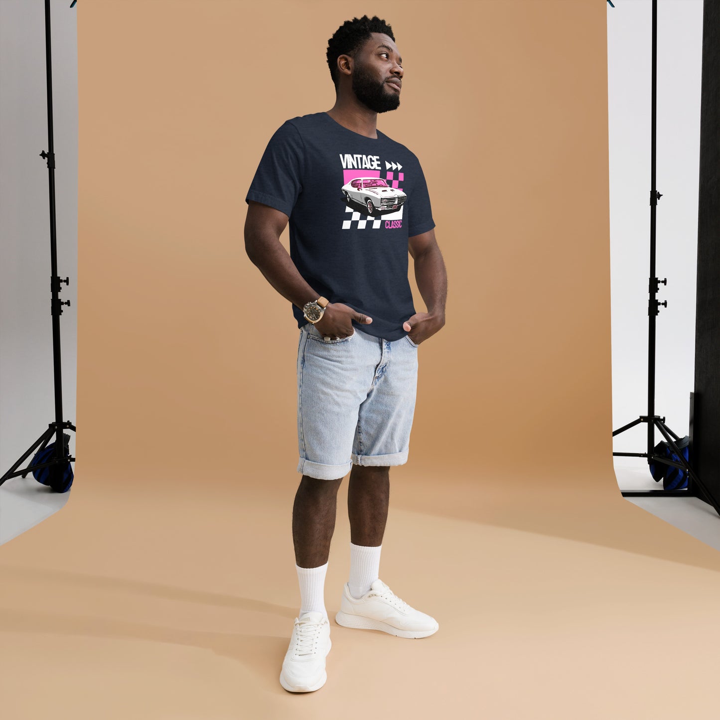 Man wearing a navy blue t-shirt with a graphic design, light blue shorts, and white sneakers in a studio setting.