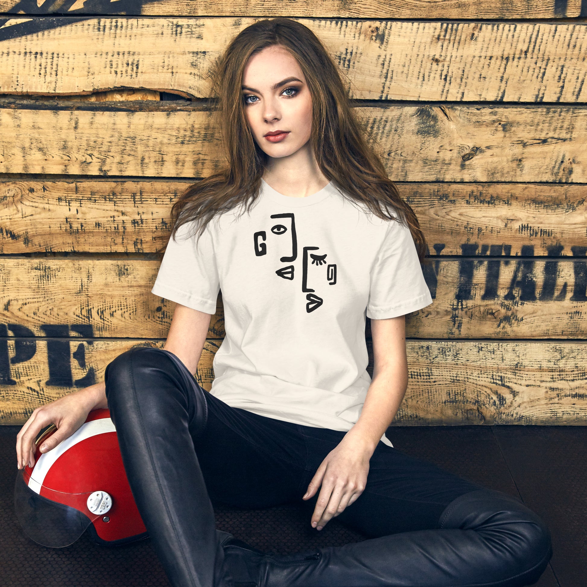 Woman wearing a white t-shirt with a graphic design, sitting against a wooden wall.